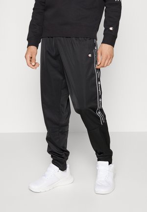Homme portant un pantalon de survêtement Champion noir avec des bandes logo sur les côtés et des baskets blanches sur un fond uni.