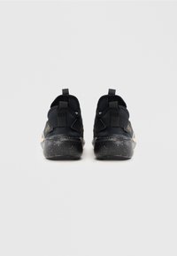 Fekete slip-on tornacipő pár speckled talppal, sarokhurokkal és arany kiegészítőkkel, hátulról, egyszínű háttér előtt.