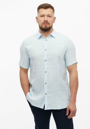 Man met baard, die een lichtblauw overhemd met korte mouwen en knopen draagt en donkere broek, staand met één hand in de zak op een witte achtergrond.