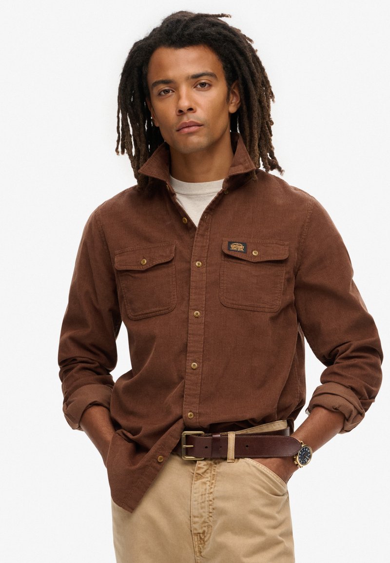 Bruine corduroy shirt met knoop-nek, twee borstzakken en gouden hardware details, gecombineerd met lichtbeige broek en een bruine riem.