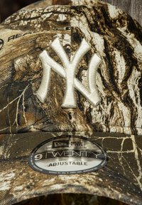 Gorra de béisbol de camuflaje con un logo de los New York Yankees bordado en relieve en color crema. La tela tiene un patrón de camuflaje woodland texturizado.