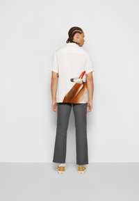 Chemise blanche à manches courtes avec un motif graphique coloré au dos, associée à un pantalon gris et des baskets jaunes.