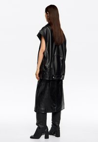 Melnas ādas veste ar plašām roku atverēm, apvienota ar saderīgu melnu ādas svārku. Aizmugures skats parāda gludu tekstūru un pieguļošu siluetu.