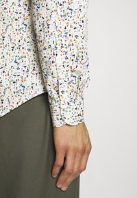 Chemise blanche à manches longues avec de petites taches multicolores ressemblant à de la peinture, portée avec un pantalon vert foncé, montrant le poignet et la main sur le côté.