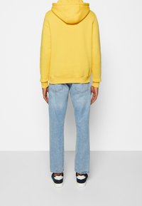 Sudadera con capucha amarilla, jeans de mezclilla azul y zapatillas azul marino. La sudadera tiene un bolsillo tipo canguro y puños acanalados. Corte holgado.