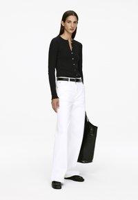 Schwarzes Langarmhemd mit Knöpfen, weiße weit geschnittene Jeans, schwarzer Gürtel und schwarze Tote-Tasche. Das Model steht und trägt schwarze flache Schuhe.