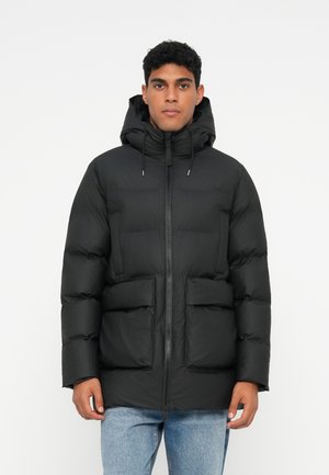 Uomo che indossa un giubbotto puffer nero con cappuccio e tasche frontali, insieme a jeans blu, in piedi di fronte a uno sfondo bianco semplice.