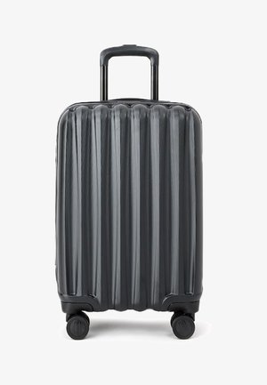 Czarna twarda walizka z pionowym ryflowanym wzorem, podwójnymi kółkami i wysuwanym uchwytem. Posiada zamek błyskawiczny.