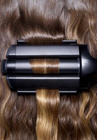 Crêpeuse à cheveux stylisant des cheveux bruns, une section lisse maintenue dans une crêpeuse à cheveux noire à triple baril chauffée pour des vagues texturées.