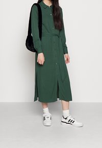 Kobieta w długiej zielonej sukience zapinanej na guziki z przewiązanym w pasie paskiem, w białych skarpetkach, białych sneakerach Adidas i czarnej, puszystej torebce na ramię.