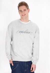 Felpa grigia con maniche lunghe e polsini a coste. Presenta la scritta "esplora all'infinito" in blu. Indossata con una t-shirt bianca sotto.