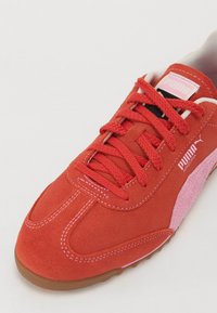 Červené semišové tenisky s růžovými akcenty, vybavené plochými tkaničkami, bílým prošitím a gumovou podešví. Logo Puma na boku a na jazyku.
