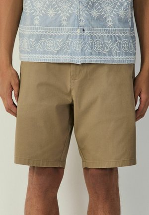 Mann trägt hellblau gemustertes Hemd und beige knielange Shorts, steht mit entspannten Händen an den Seiten.