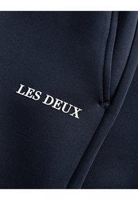 Navy athletic pants with a smooth texture and white "LES DEUX" logo. Features tapered design and visible stitching details.