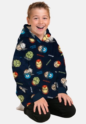 Smiling boy kneeling, wearing a navy hoodie patterned with Marvel superhero faces including Iron Man, Captain America, Hulk, and Thor.