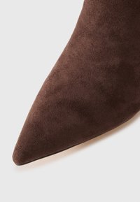 Barna velúr, hegyes orrú csizma, sima, puha textúrával és elegáns vonalvezetéssel, enyhén megemelt sarokkal a stílusos megjelenés érdekében.