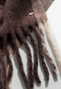 Smeđi i bež čupavi šal od vune sa mekanom teksturom, sa pletenom resicom i malom metalnom oznakom. Boja se gradiira od tamne do svijetle.