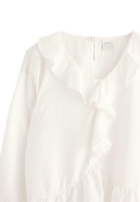 Vit långärmad blus med subtil prickig textur, volangkrage och framknäppning vid bakkant av kragen.