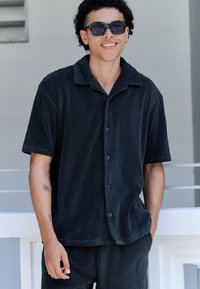 Camicia nera a maniche corte con una texture e un motivo geometrico, caratterizzata da una chiusura con bottoni sul davanti e un tessuto morbido e vellutato. Sono visibili anche pantaloncini coordinati.