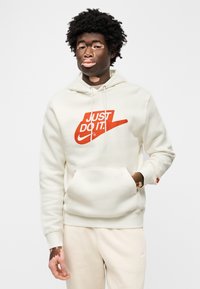 Λευκό φούτερ με κουκούλα και εμφανές κόκκινο λογότυπο "JUST DO IT.", με μπροστινή τσέπη και κορδόνι στην κουκούλα, συνδυασμένο με ανοιχτόχρωμο παντελόνι.
