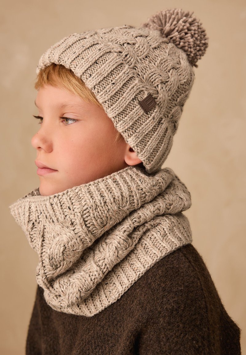Enfant portant un bonnet beige en tricot avec un pompon et une écharpe assortie épaisse, ainsi qu'un pull marron foncé, regardant vers la gauche sur un fond neutre.