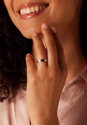 Bague en argent avec une surface lisse et polie portée sur une main touchant le visage, mettant en valeur un design moderne et subtil contre une blouse claire.
