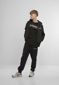 Black zip-up hoodie with logo, black t-shirt with white text, black sweatpants with logo, and metallic-patterned running shoes.