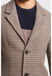 Hombre con un blazer a rayas marrón, negro y blanco sobre un suéter de punto azul marino con un panel central texturizado.