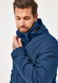 Mann in einer blauen Kapuzenjacke, der mit einer tätowierten Hand den Kragen anpasst und nach unten schaut, vor einem schlichten Hintergrund.