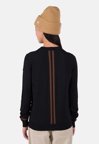 Schwarzer Pullover mit gerippten Bündchen und Saum, der braune vertikale Streifen auf dem Rücken hat. Getragen mit einer beige gestrickten Mütze.