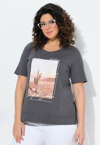 Grijze T-shirt met korte mouwen met een afbeelding aan de voorkant van een woestijnlandschap met een cactus, zonnestralen en de tekst "Eindeloze Dagen van de Zomer."