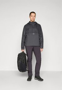 Femme en veste noire et pantalons sombres tenant un sac à dos noir, se tenant devant un fond gris clair uni.