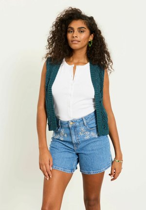 Žena s kudrnatými vlasy, která má na sobě zelenou pletenou vestu, bílý sleeveless top na knoflíky, modré džínové šortky s výšivkou a zelené náušnice, stojí.