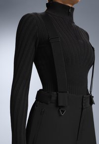 Černý žebrovaný top s dlouhými rukávy, vysokým límcem a zipem, v kombinaci s černými kšandami na hladkých, strukturovaných kalhotách s kapsami na zip.