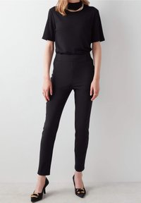 Osoba v černém krátkém tričku a úzkých černých kalhotách stojí proti světlému šedému pozadí, doplněná černými podpatky.