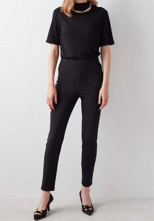 Iemand in een zwart t-shirt met korte mouwen en slanke zwarte broeken staat tegen een lichtgrijze achtergrond, aangevuld met zwarte hakken.