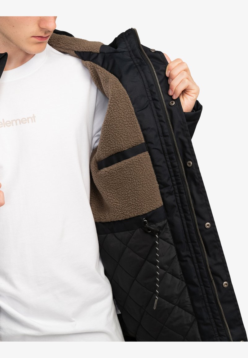 Chaqueta aislante negra con un diseño acolchado, forro de forro polar marrón y cierres de botones a presión. Presenta bolsillos interiores y un cordón ajustable.