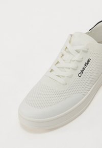 Adidași albi din plasă, cu șireturi plate, vârful rotunjit, panouri laterale texturate și un mic logo negru pe partea exterioară. Talpă din cauciuc neted.