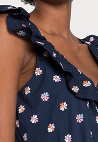 Marinblå blommig klänning med rynkad halsringning och ärmlös design. Tyget har små, flerfärgade blommönster över hela klänningen.