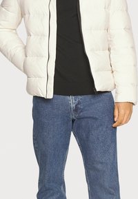 Helle, gep puffte Jacke mit Reißverschluss, die ein gestepptes Design aufweist. Kombiniert mit einem dunklen Hemd und blauen Denim-Jeans.