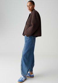 Giacca marrone oversize con finitura testurizzata, abbinata a jeans denim blu larghi e sneakers blu con strisce bianche.