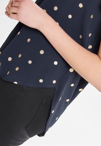 Mørkeblå top med gyldne prikker, lavet af letvægtsstof, med en buet søm og en afslappet pasform. Sammen med sorte bukser.