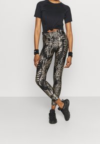 Top nero a maniche corte annodato in vita, abbinato a leggings a vita alta con motivo astratto nero e oro metallizzato. Scarpe da ginnastica nere.