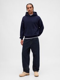 Bărbat cu păr împletit, îmbrăcat într-o hanorac albastru închis, blugi negri largi și cizme bej cu șireturi, stând pe un fundal alb simplu.