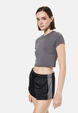 Jonge vrouw draagt een aansluitend grijs cropped T-shirt en zwarte sportshorts met een grijze zijstreep, lichtjes opzij kijkend tegen een witte achtergrond.