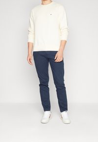 Sudadera crema con puños y dobladillo acanalados, combinada con jeans navy y zapatillas blancas con detalles rojos. Conjunto simple y casual.