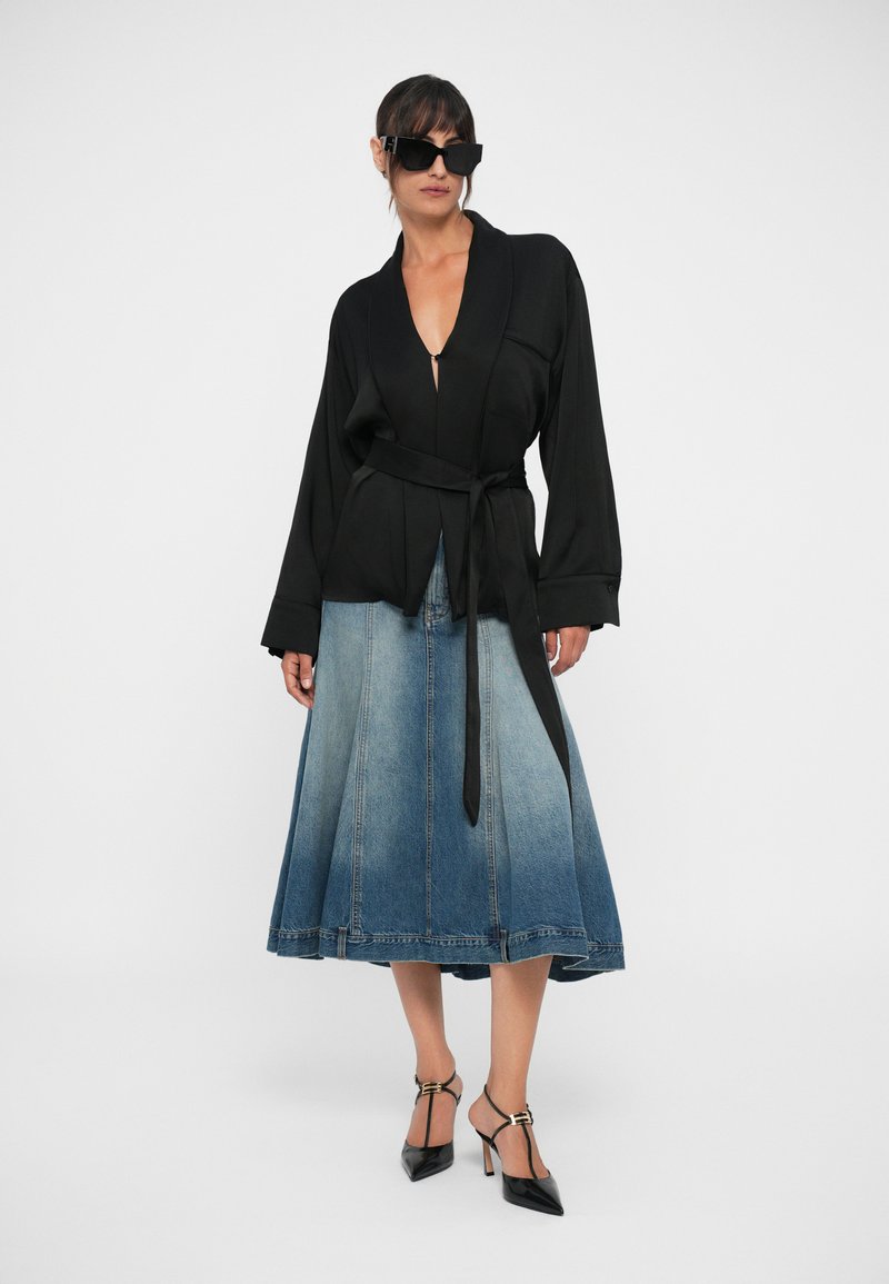 Schwarze Wickelbluse mit langen Ärmeln, kombiniert mit einem Denim-Rock im Ombre-Effekt. Die Accessoires umfassen schwarze Sonnenbrillen und Absatzschuhe.