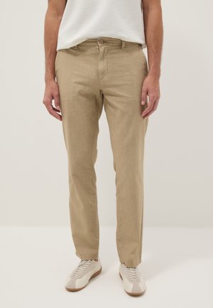 Uomo che indossa pantaloni beige dritti, sneakers bianche e una maglietta bianca a maniche corte con texture, in piedi contro uno sfondo chiaro e uniforme.