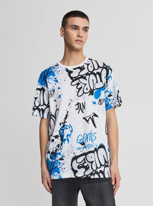 Giovane uomo che indossa una maglietta bianca oversize con testo in stile graffiti blu e nero e motivi a schizzi di vernice, in piedi davanti a uno sfondo semplice.