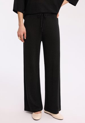 Czarne spodnie o szerokich nogawkach z wiązanym paskiem w talii, noszone z beżowymi balerinami i czarnym topem, ręka spoczywająca na biodrze.